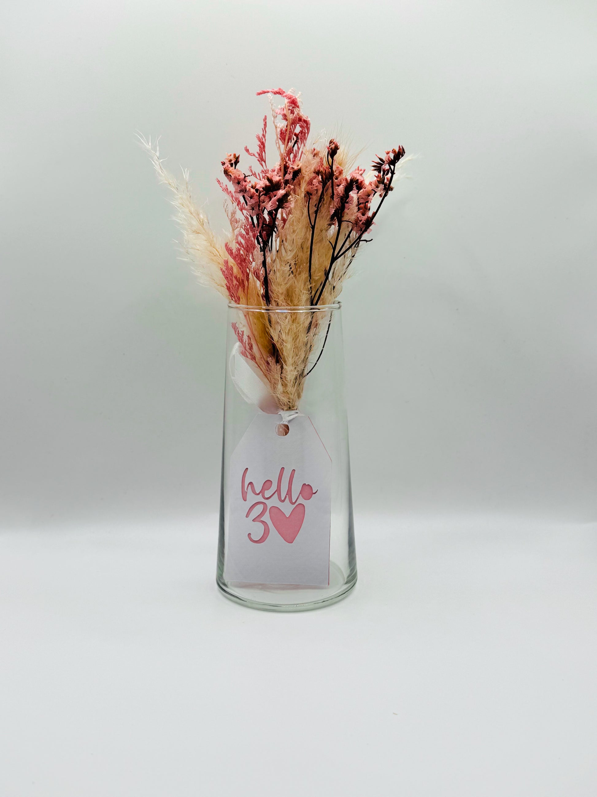 Glass vase with dried flowers and a 'hello 3' tag on a white background